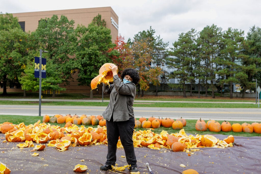A person wearing a mask is lifting a carved pumpkin, surrounded by pumpkin remnants and whole pumpkins on a grassy area.