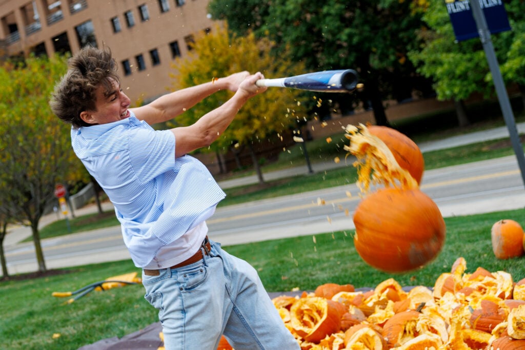 A young man swings a baseball bat to hit a pumpkin, causing pumpkin guts to fly in the air, with many smashed pumpkins in the background.