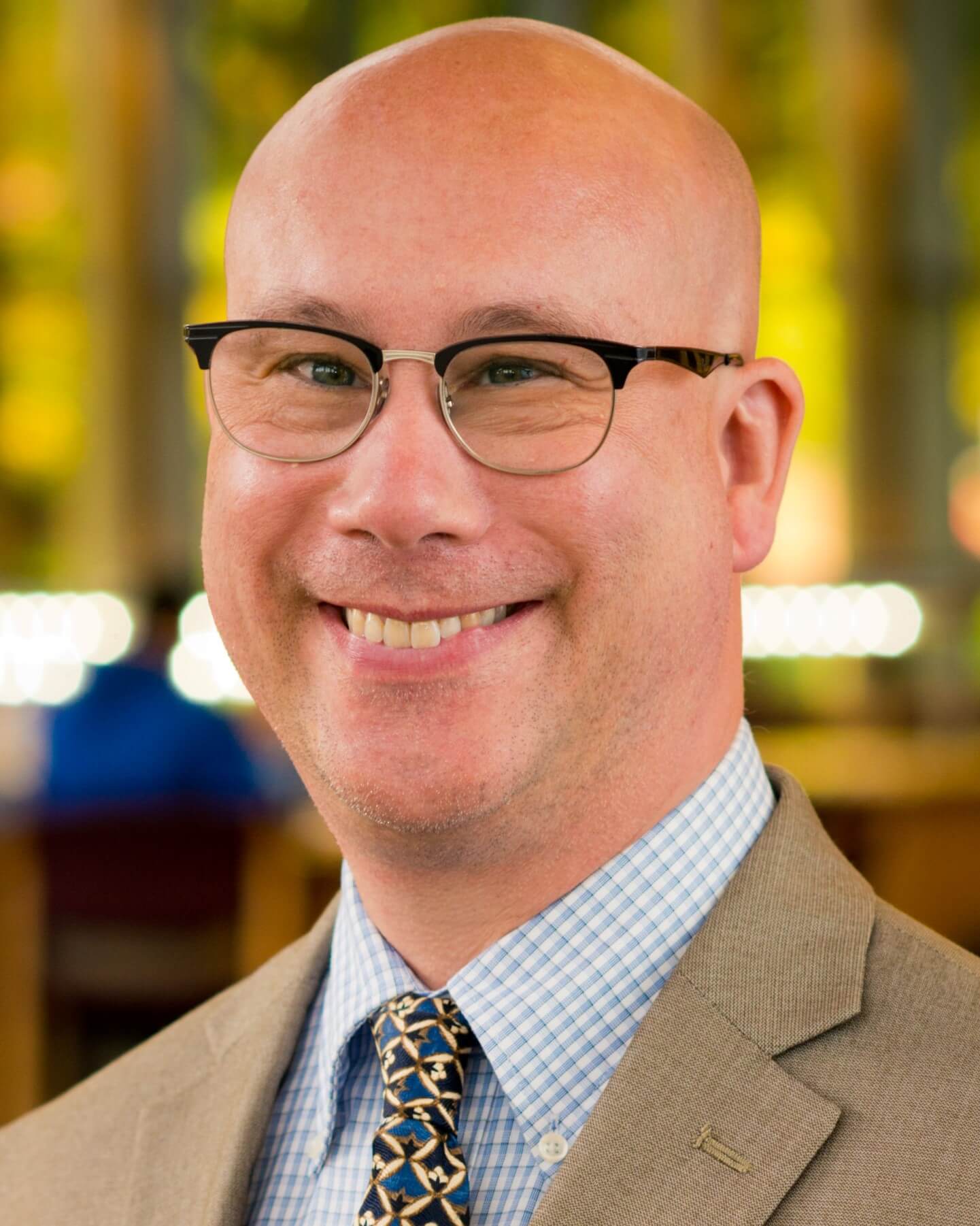 A professional portrait of a smiling bald man. He is wearing glasses, a light-brown suit, and a blue, checkered shirt with a patterned tie.