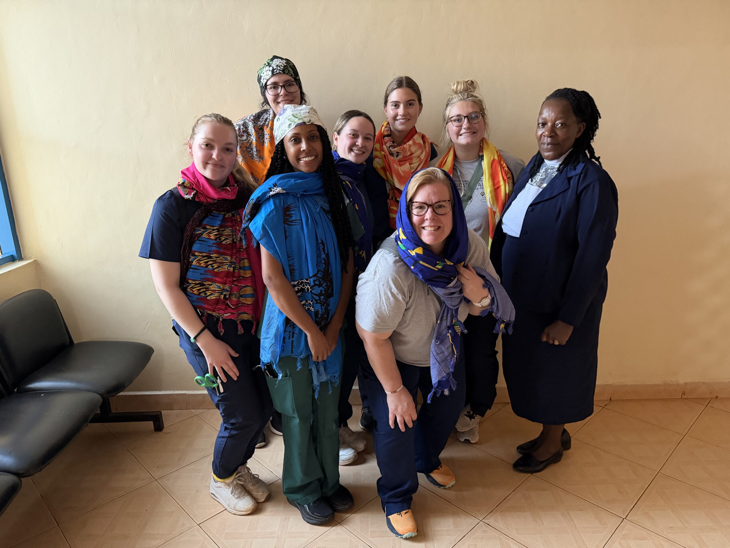 A group of eight women, some wearing colorful scarves, pose together in a room with a light-colored wall and a bench.