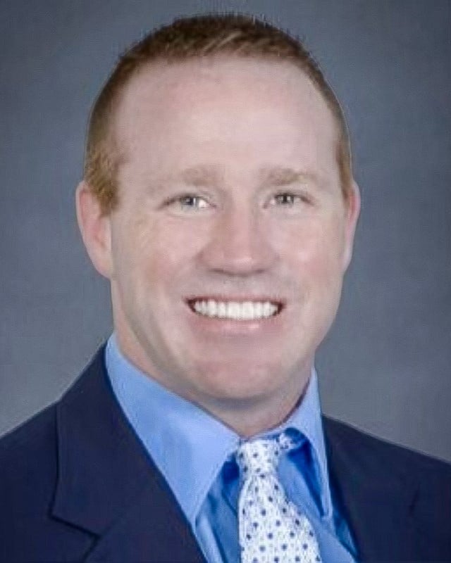 A professional portrait of a man with short red hair wearing a blue business suit and patterned white tie.