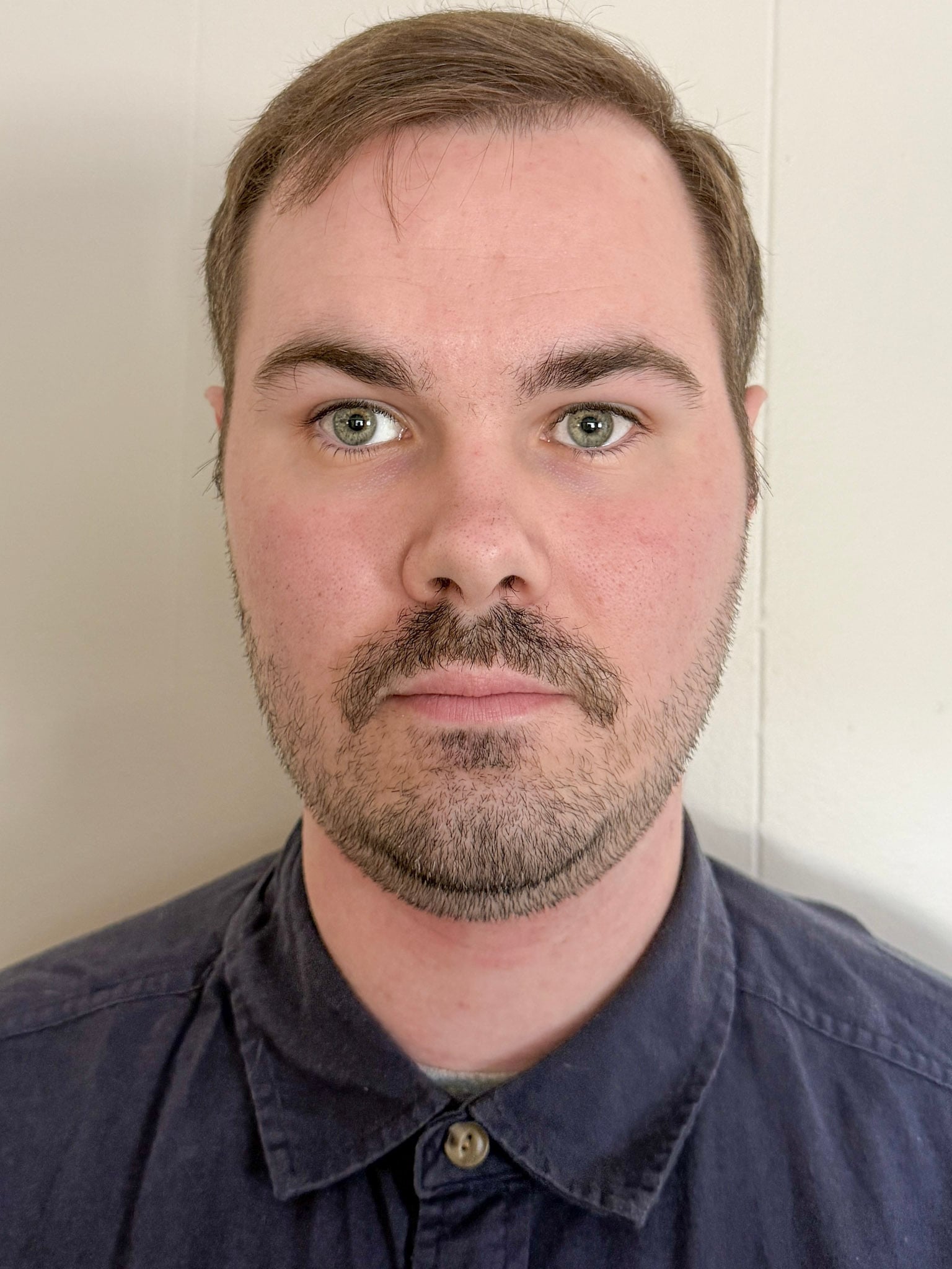 A close-up portrait of a man with short brown hair and a short beard. He is wearing a navy-blue collared shirt against a light background.