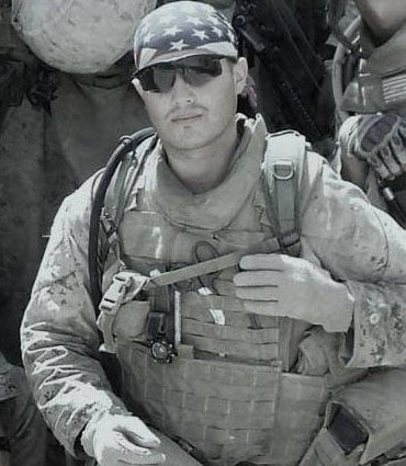 A soldier wearing sunglasses and a bandana with an American flag, dressed in military gear, poses while crouching.