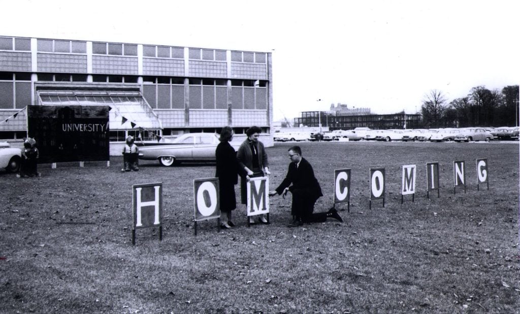 Black and white image of staff putting up a homecoming yard sign on a lawn in front of a building.