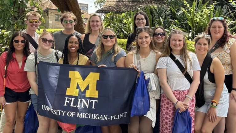 A group of people pose outdoors, holding a flag that reads "M FLINT UNIVERSITY OF MICHIGAN-FLINT." They are smiling and wearing casual clothing, with greenery and a large sculpture in the background.