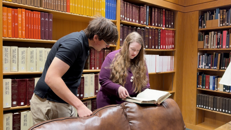 Two people are looking at a large book while seated on a couch in a library, surrounded by shelves of books.