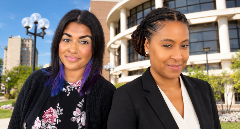 Two women stand outdoors in front of a modern building. One has long, dark hair with purple tips and wears a floral top, while the other has short, styled hair and is dressed in a black suit.