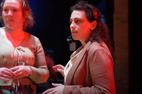 Two women are engaged in conversation in a dimly lit setting. One woman, wearing a headset and a blazer, appears to be giving instructions or guidance. The other woman, in a light green top, listens attentively.