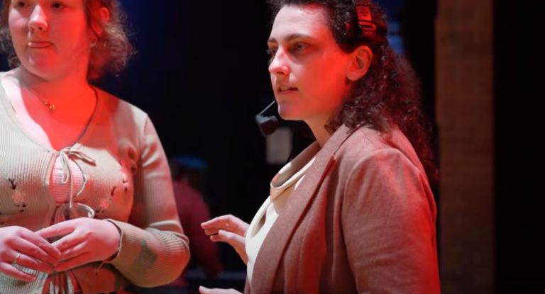 Two women are engaged in conversation in a dimly lit setting. One woman, wearing a headset and a blazer, appears to be giving instructions or guidance. The other woman, in a light green top, listens attentively.