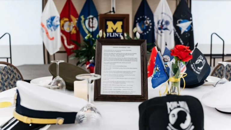 A table set for an event featuring military hats, a red rose in a vase, a framed document, and various flags in the background.