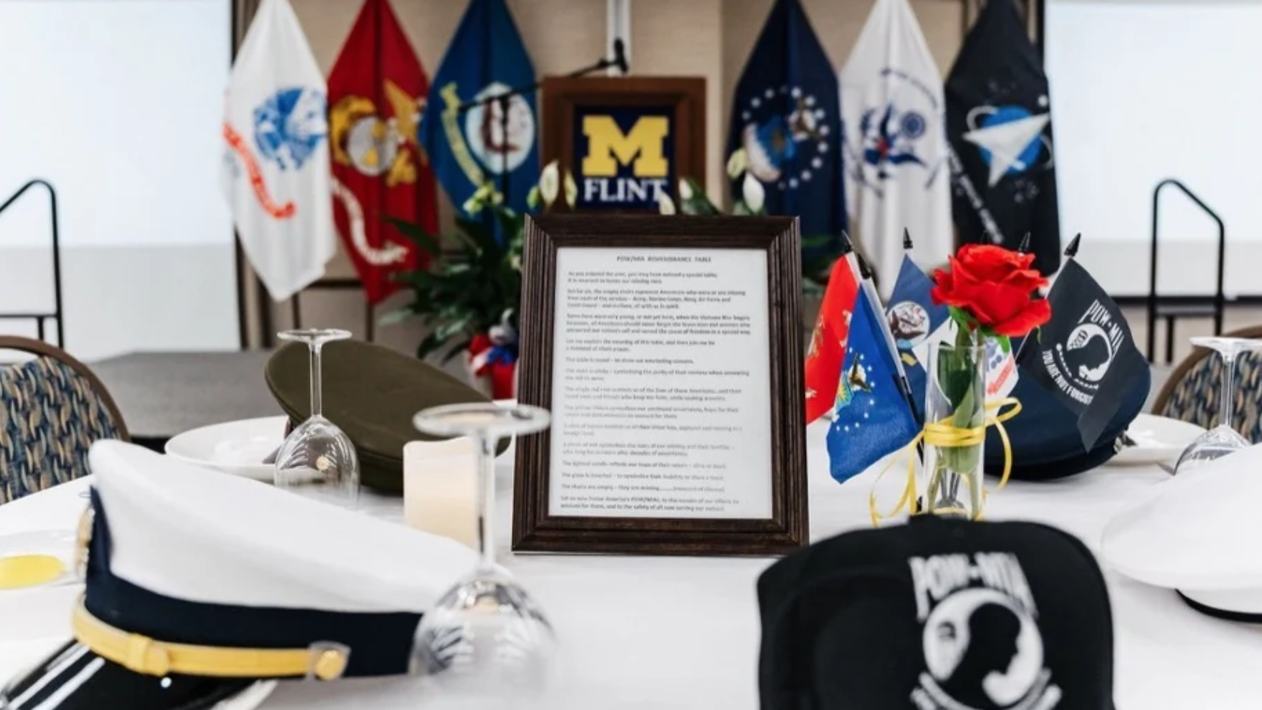 A table set for an event featuring military hats, a red rose in a vase, a framed document, and various flags in the background.